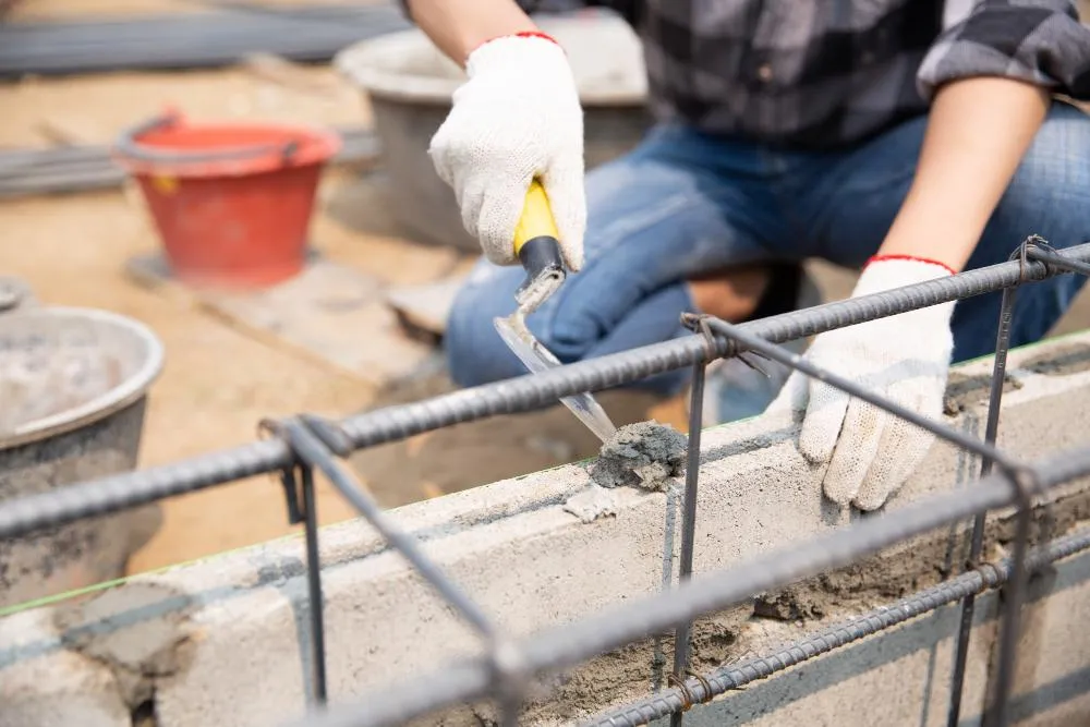 Construction worker wearing gloves uses a trowel to apply cement on a reinforced concrete wall with steel rebar framework.