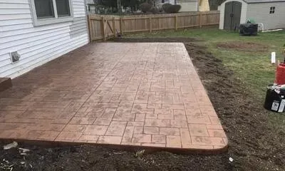 A newly installed decorative stamped concrete patio beside a house, featuring a brick-patterned texture and surrounded by a fenced backyard.
