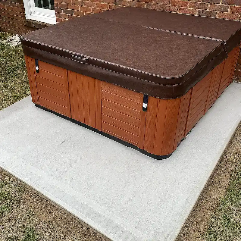 Hot tub on concrete slab with wooden sides and brown colored cover next to a brick wall