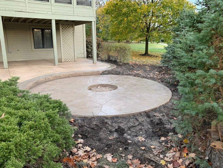 A freshly built circular concrete patio with a center cut-out for a fire pit, located in a backyard surrounded by trees and shrubs.
